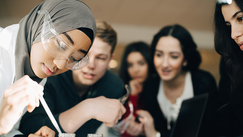 Teacher demonstrating an experiment with teenage students standing in a group